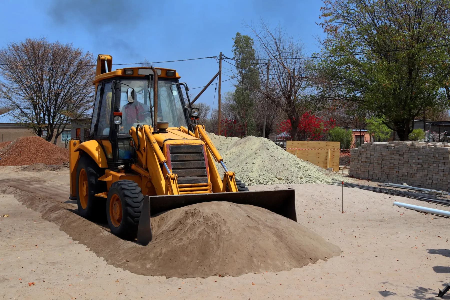 A yellow TLB pushing a pile of sand on a building site, with stacked bricks and trees behind